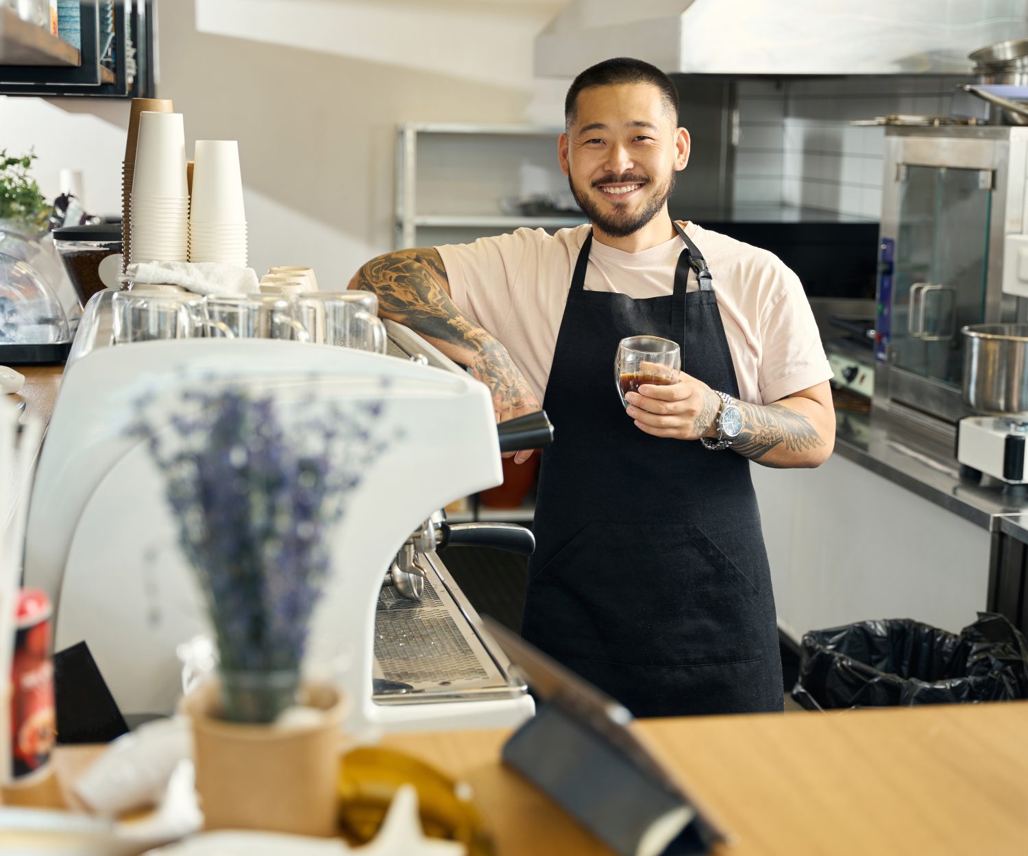 Cheerful young barista smiling widely and holding a cup of espresso while posing at a coffee shop